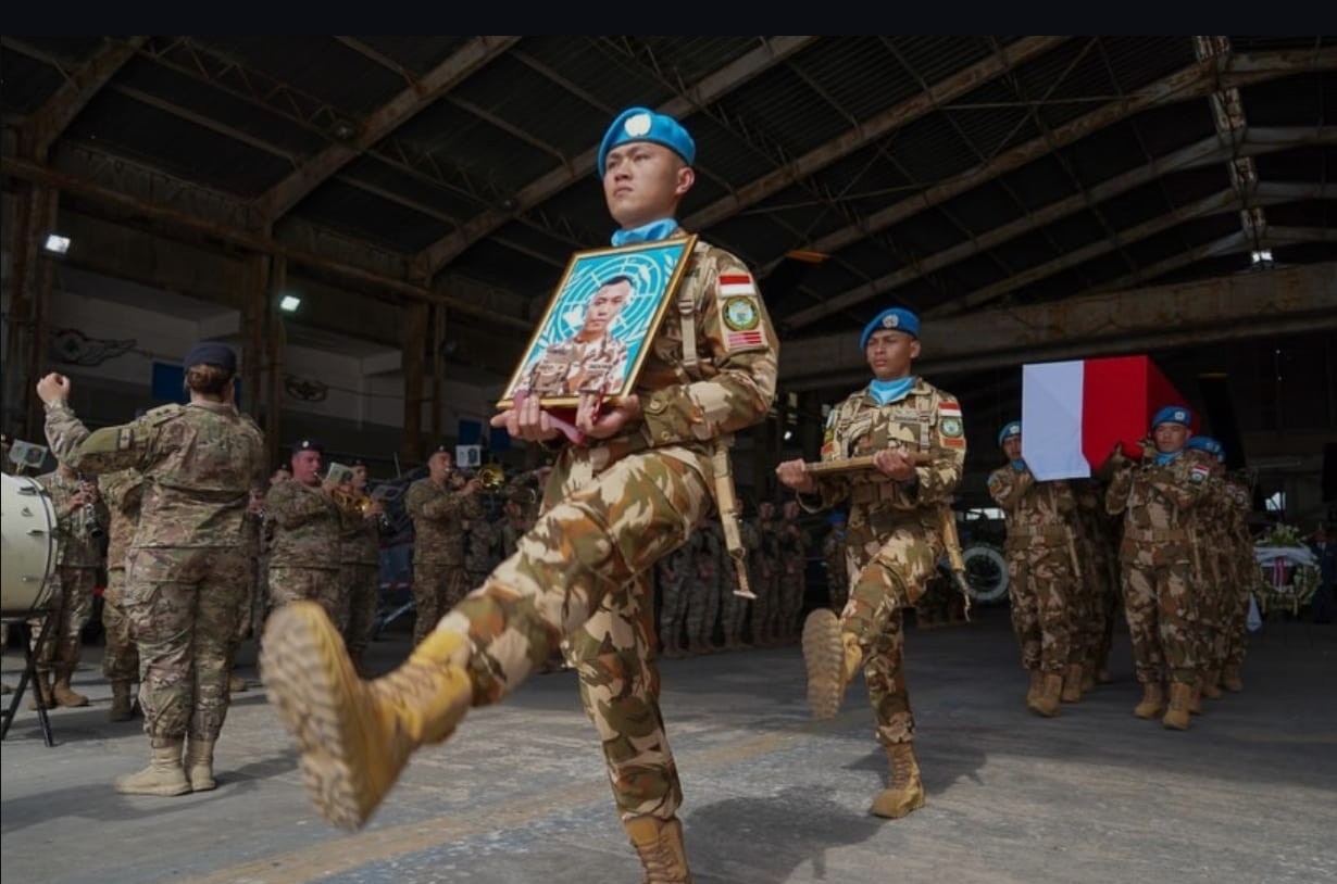 Soldiers in camouflage march inside a hangar the lead soldier carries a framed portrait of a fallen comrade while others follow with a flag draped coffin in the background