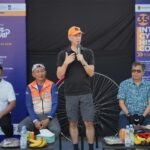 Man in black polo speaking into a microphone on stage flanked by purple event banners and seated colleagues fruit and drinks on tables in front