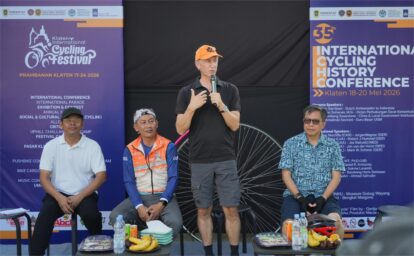 Man in black polo speaking into a microphone on stage, flanked by purple event banners and seated colleagues; fruit and drinks on tables in front.