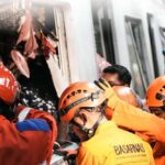 Emergency responders in orange uniforms and helmets extract passengers from a damaged train car amid debris during a rescue operation