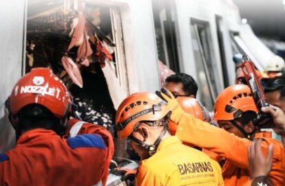 Emergency responders in orange uniforms and helmets extract passengers from a damaged train car amid debris, during a rescue operation.