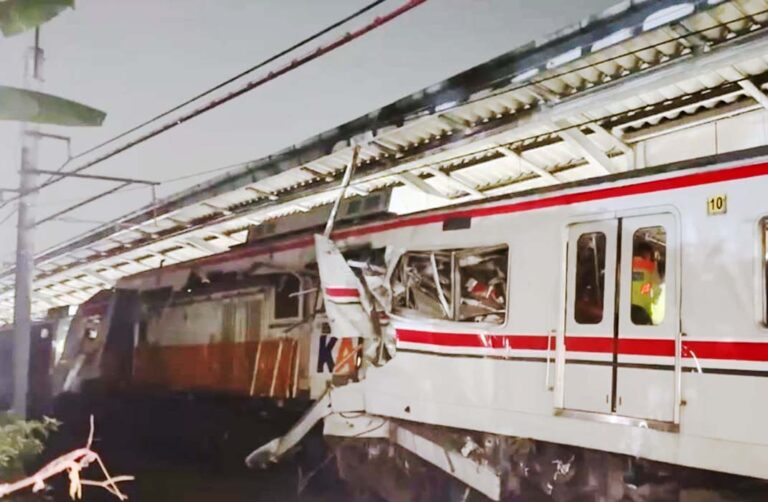 White commuter train with red stripes derailed against a platform, front area severely damaged with bent metal and shattered windows.