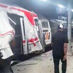 Damaged passenger train at a station platform with doors open and debris on the floor a man stands nearby as an emergency vehicle is visible in the background