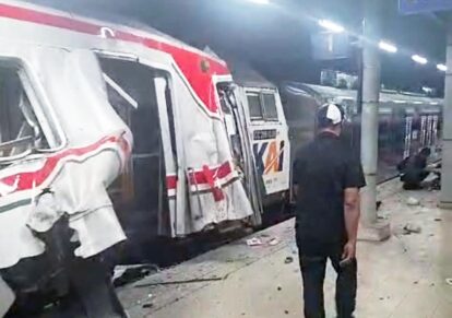Damaged passenger train at a station platform with doors open and debris on the floor a man stands nearby as an emergency vehicle is visible in the background