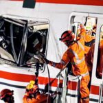 Emergency responders in orange gear perform a rescue from a damaged rail vehicle crash site extricating occupants from a smashed train car
