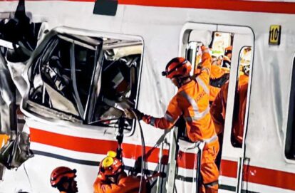 Emergency responders in orange gear perform a rescue from a damaged rail vehicle crash site extricating occupants from a smashed train car
