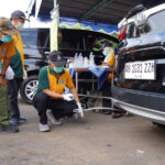 A team of workers in green and yellow uniforms and masks perform a car diagnostic under a tent one person kneels to connect cables to the rear of a dark suv while others watch nearby