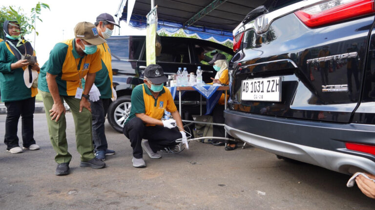 A team of workers in green and yellow uniforms and masks perform a car diagnostic under a tent; one person kneels to connect cables to the rear of a dark SUV while others watch nearby.