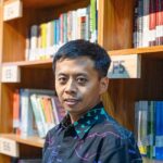 Man standing in a library or bookstore aisle looking at the camera with shelves of colorful books behind him