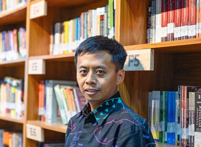 Man standing in a library or bookstore aisle, looking at the camera with shelves of colorful books behind him.