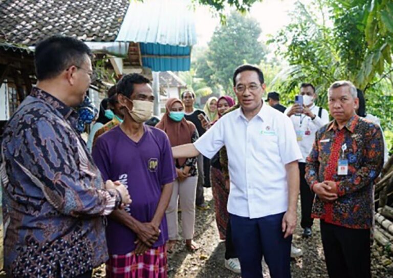 Man in white shirt shakes hands with a man in a purple shirt as a small crowd watches outdoors, in a rural setting.