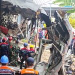 Emergency responders in helmets and uniforms inspect a collapsed wreckage with tangled metal and debris scattered along a ramp or street