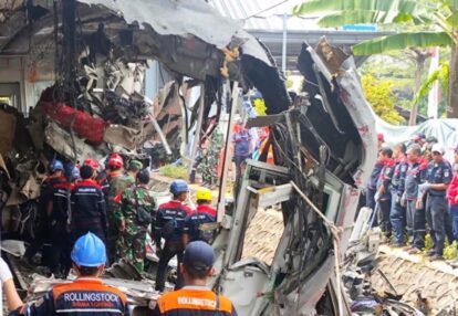 Emergency responders in helmets and uniforms inspect a collapsed wreckage with tangled metal and debris scattered along a ramp or street.
