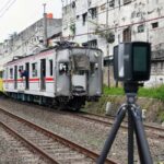 Damaged commuter train on tracks with peeling buildings in a city and a camera on a tripod in the foreground used for surveying