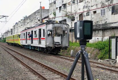 Damaged commuter train on tracks with peeling buildings in a city, and a camera on a tripod in the foreground used for surveying.