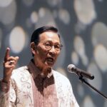 Man in a patterned batik shirt speaks into a microphone raising his index finger as he makes a point