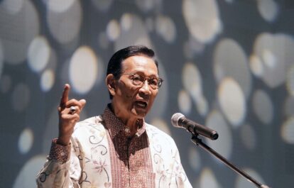 Man in a patterned batik shirt speaks into a microphone raising his index finger as he makes a point