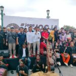 Large group of people on a stage posing for a photo behind a white banner with jogja text and circular logo hints outdoors at event