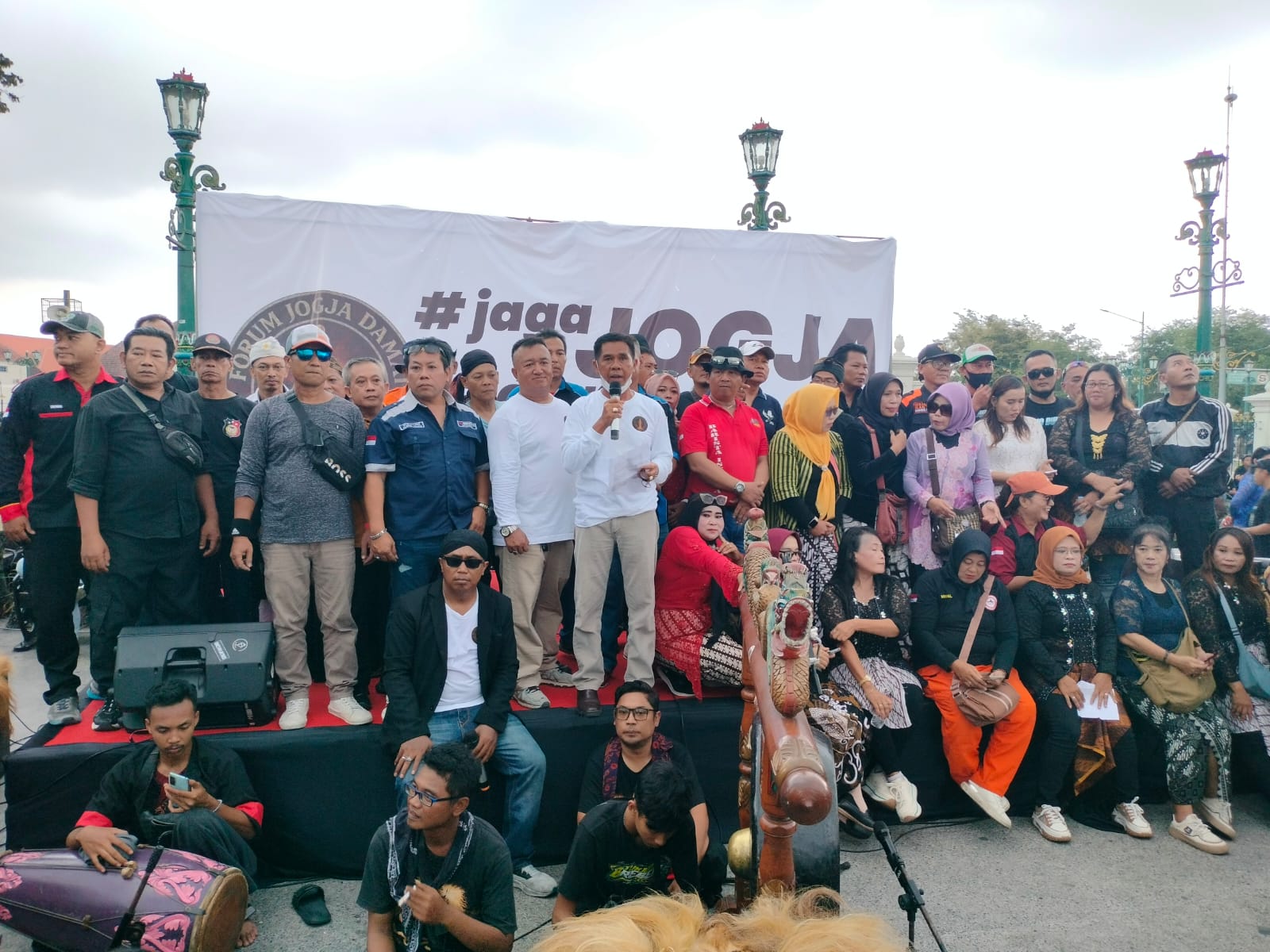 Large group of people on a stage posing for a photo behind a white banner with jogja text and circular logo hints outdoors at event