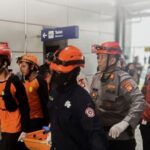 Emergency responders in orange vests and helmets escorting a person on a wheeled stretcher through a building corridor