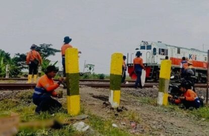 Railroad workers in orange uniforms kneeling and painting yellow posts along the track with a train in the background.