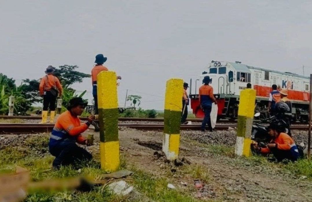 Railroad workers in orange uniforms kneeling and painting yellow posts along the track with a train in the background