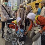 Uniformed volunteers assist an elderly woman in a wheelchair on a city sidewalk beside a bright yellow bus