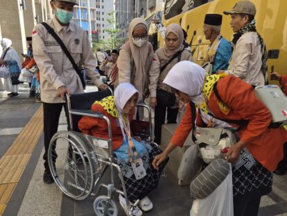 Uniformed volunteers assist an elderly woman in a wheelchair on a city sidewalk beside a bright yellow bus
