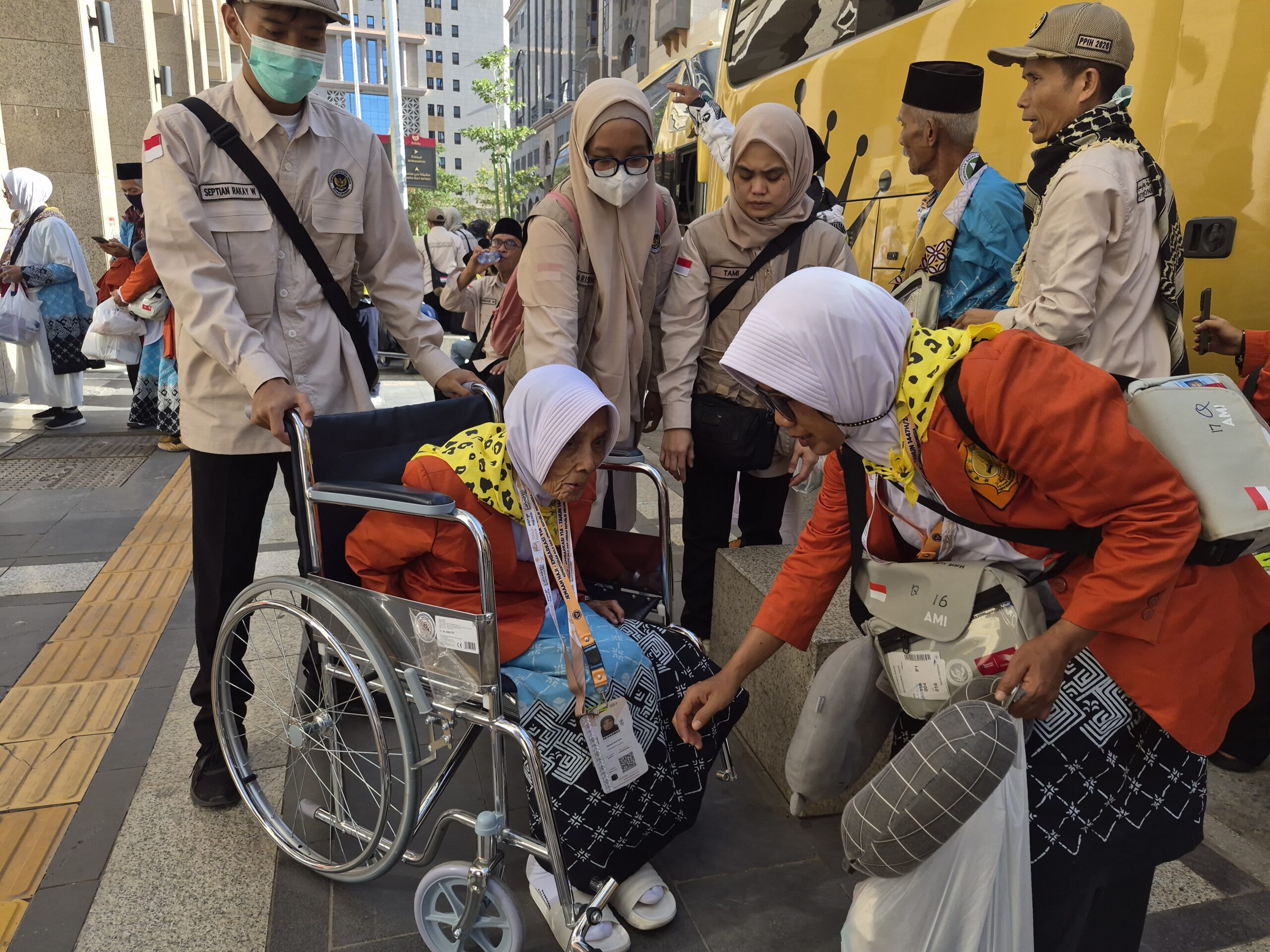 Uniformed volunteers assist an elderly woman in a wheelchair on a city sidewalk beside a bright yellow bus