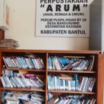 A cluttered wooden bookshelf filled with magazines and papers beneath a large sign reading perpustakaan arum anak remaja  umum in indonesian