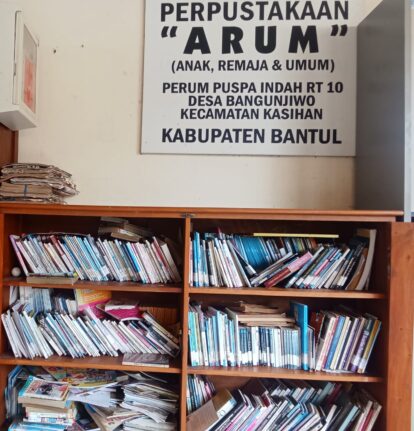 A cluttered wooden bookshelf filled with magazines and papers beneath a large sign reading 'PERPUSTAKAAN ARUM (ANAK, REMAJA & UMUM)' in Indonesian.