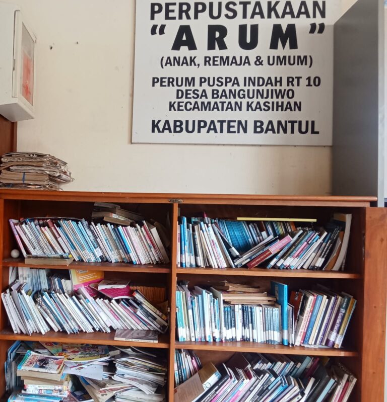 A cluttered wooden bookshelf filled with magazines and papers beneath a large sign reading 'PERPUSTAKAAN ARUM (ANAK, REMAJA & UMUM)' in Indonesian.