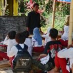 Teacher standing at front of outdoor classroom with students seated on benches and wooden tables a chalkboard behind them and tropical plants nearby