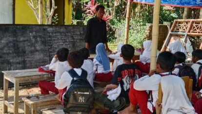 Teacher standing at front of outdoor classroom with students seated on benches and wooden tables, a chalkboard behind them and tropical plants nearby.