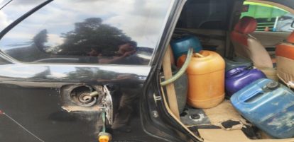 Open car door revealing several plastic gasoline containers (orange, purple, blue) occupying the back seat area; the fuel filler is exposed on the side of the car.