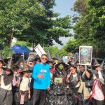A group of people wearing witch hats and dark costumes parade down a sunny street led by a man in a bright blue shirt holding a sign about environmental earth care