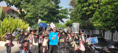 A group of people wearing witch hats and dark costumes parade down a sunny street, led by a man in a bright blue shirt, holding a sign about environmental earth care.
