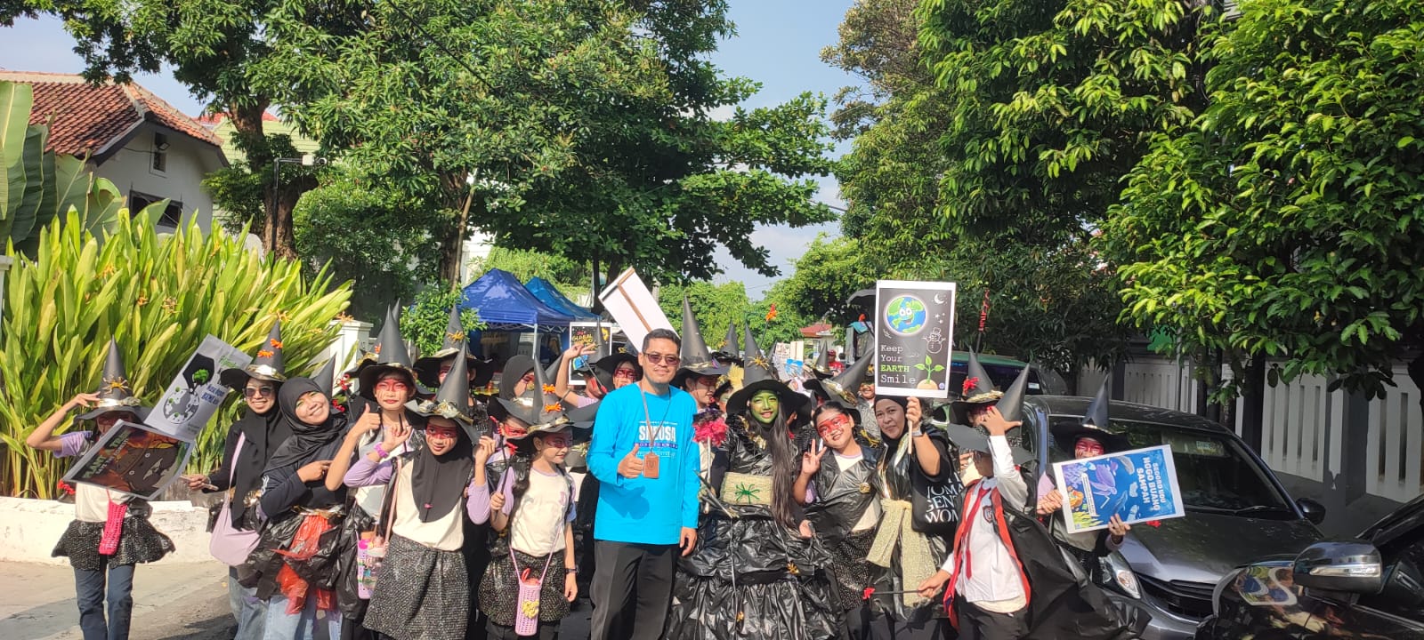 A group of people wearing witch hats and dark costumes parade down a sunny street led by a man in a bright blue shirt holding a sign about environmental earth care