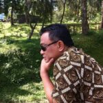 Man wearing sunglasses sits outdoors in a grassy wooded area resting his chin on his hand while looking downward