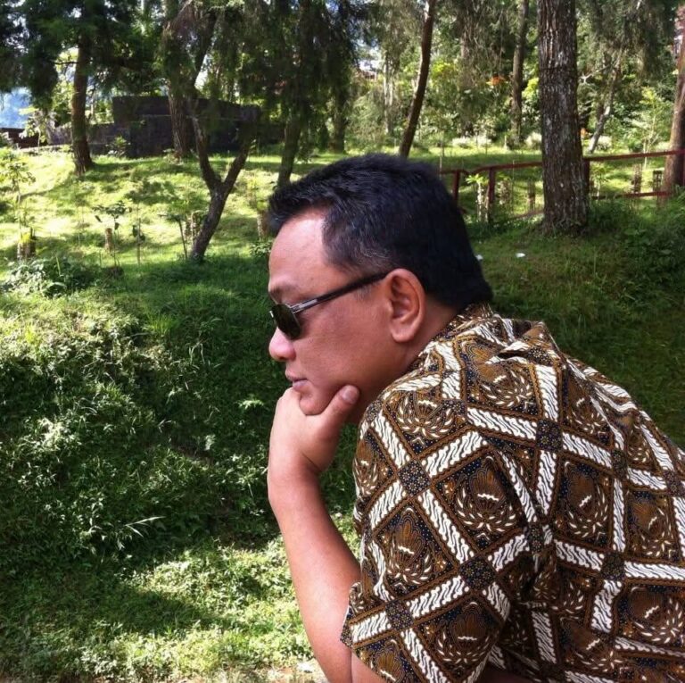 Man wearing sunglasses sits outdoors in a grassy, wooded area, resting his chin on his hand while looking downward.