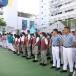 Students in uniforms standing in neat rows for a school assembly on a green outdoor court with colorful tents in the background