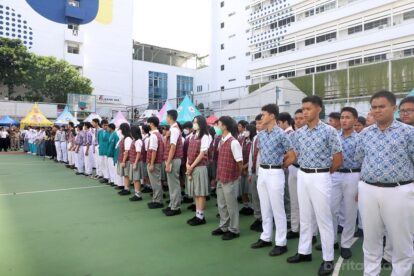 Students in uniforms standing in neat rows for a school assembly on a green outdoor court with colorful tents in the background.