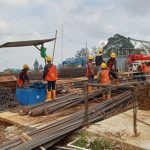 Construction site with workers in orange vests and yellow helmets arranging steel rebar and equipment on a dirt foundation blue toolbox nearby
