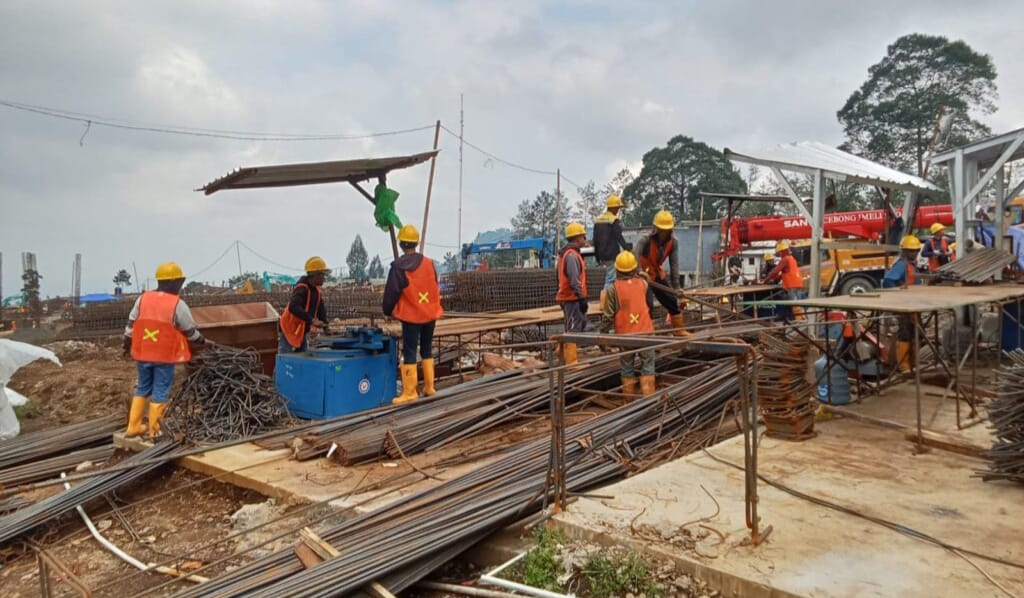 Construction site with workers in orange vests and yellow helmets arranging steel rebar and equipment on a dirt foundation blue toolbox nearby