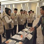 A man in batik shirt explains a display table to a line of uniformed staff wearing beige vests and black hats at a conference hall