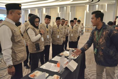 A man in batik shirt explains a display table to a line of uniformed staff wearing beige vests and black hats at a conference hall.