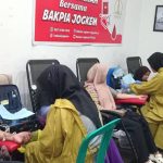Man donating blood as a nurse in a yellow uniform attends to him other donors sit in chairs in a clinic room with a red armrest