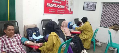 Man donating blood as a nurse in a yellow uniform attends to him other donors sit in chairs in a clinic room with a red armrest