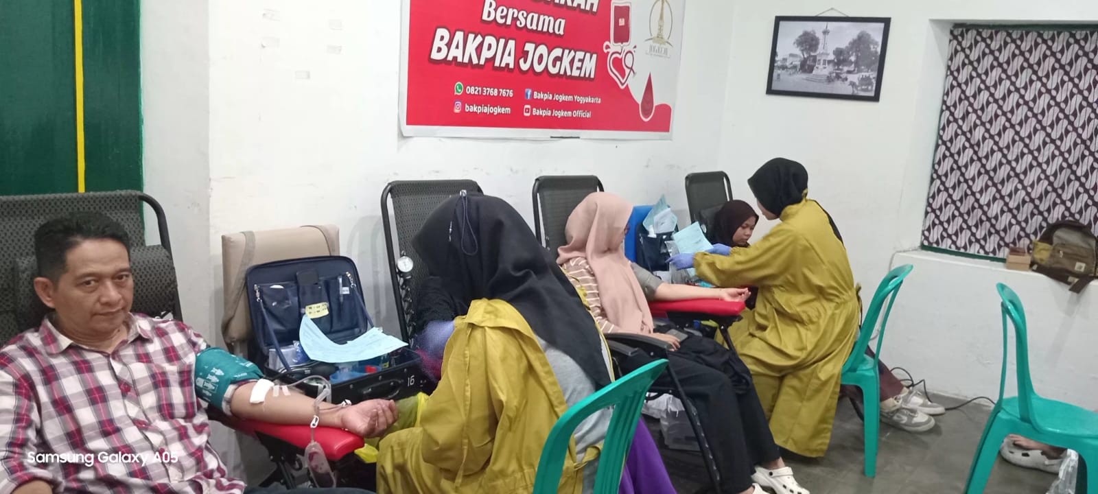 Man donating blood as a nurse in a yellow uniform attends to him other donors sit in chairs in a clinic room with a red armrest