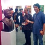 Group of men in a gallery looking at a dark bust on a white pedestal against a pink wall as they chat nearby